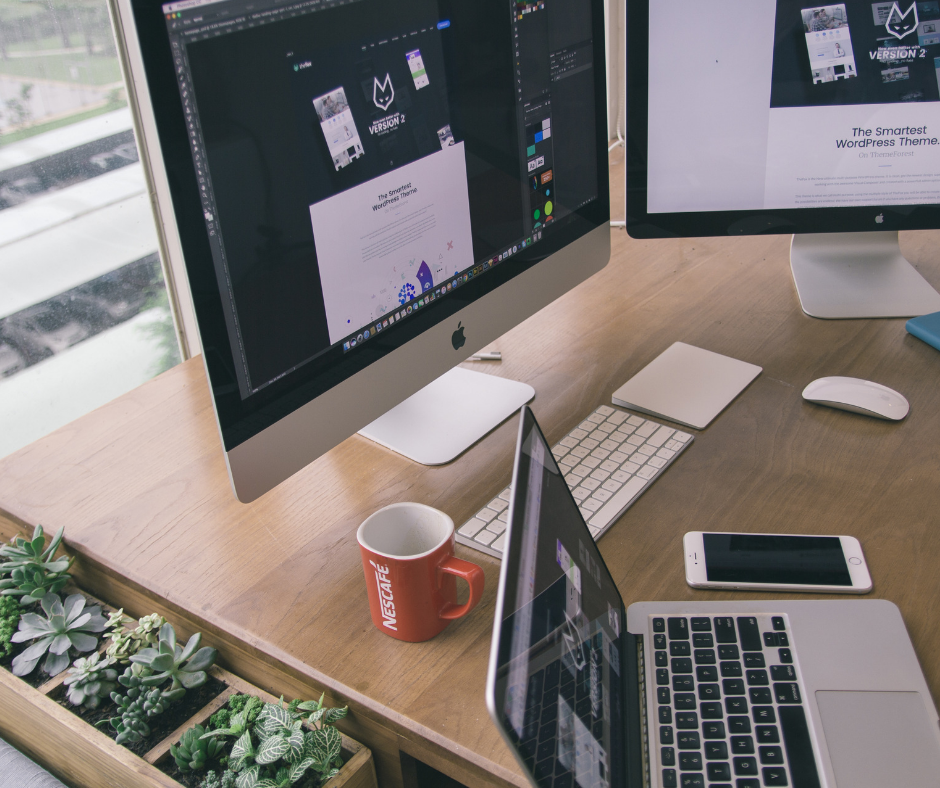 A desk by a window with various laptops and computers, a keyboard, a mouse, a phone and a mug on it, as well as a plant tray to the side of it.