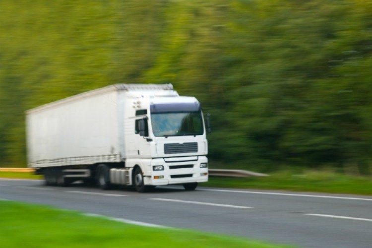 A white HGV driving down a road alongside trees and grass, it is blurred slightly to indicate its speed