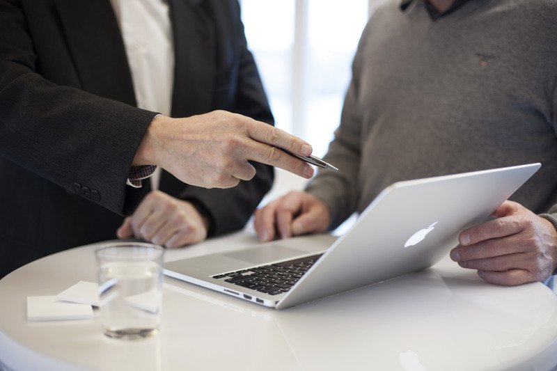 Two people conversing, one wearing a black and white suit holding a pen and the other in a grey jumper. They are looking at the screen of a Macbook which is in the middle of a white circular table with sticky notes and a glass of water on.