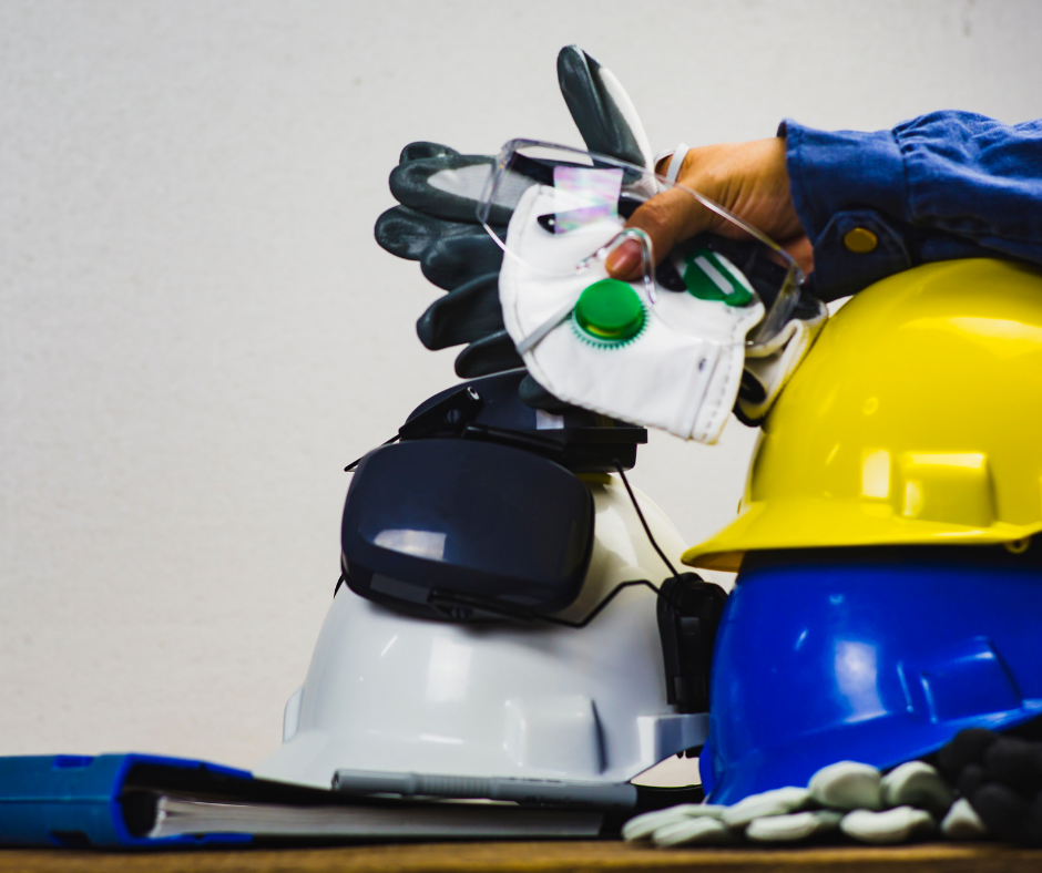 Hand holding several types of PPE including hard hats, gloves and glasses